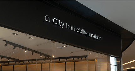 Beleuchtetes Wandschild 'City Immobilienmakler' über einer Glasfront im Innenbereich