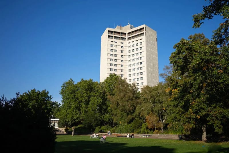 Weißer Hochhausturm hinter Bäumen mit Parkbesuchern im Vordergrund